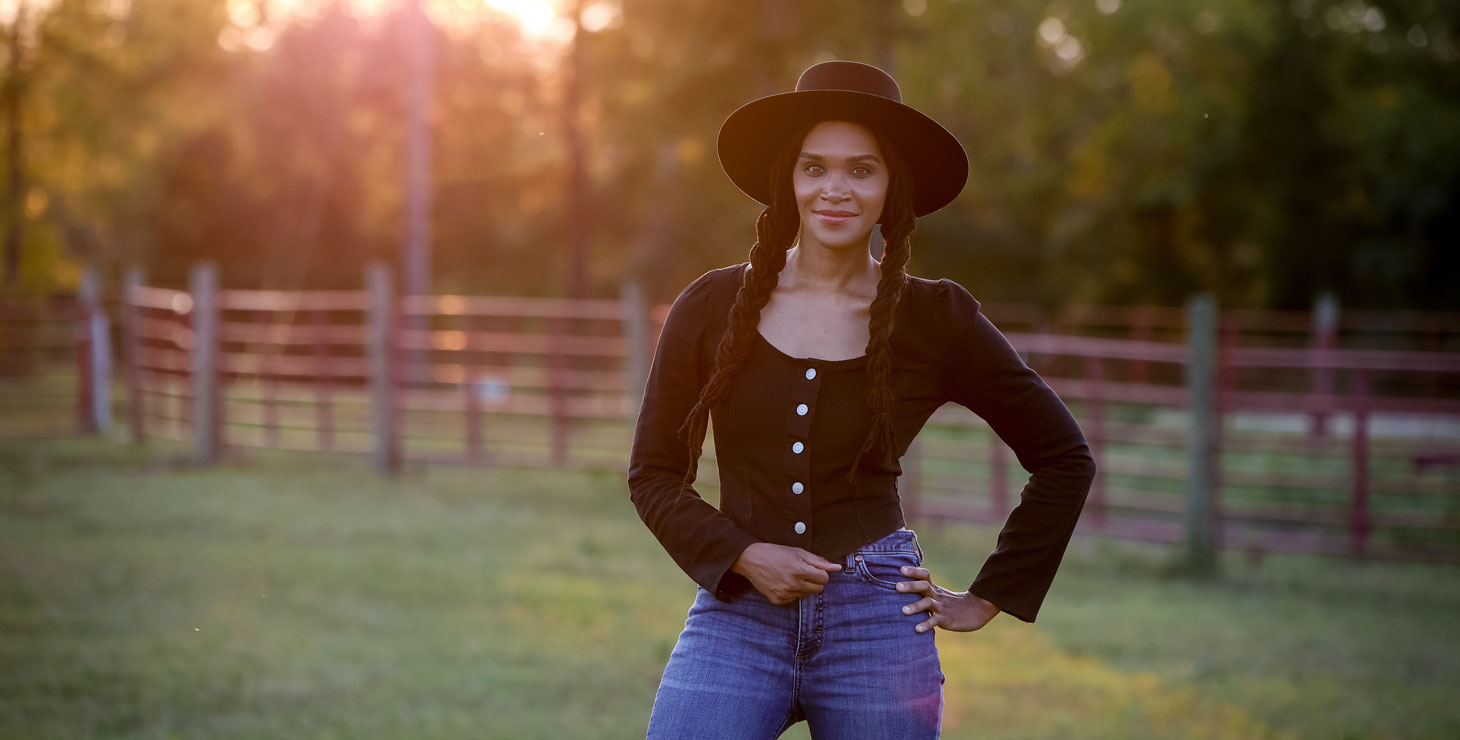 A photo of a woman wearing a black shirt, black felt hat, and blue jeans, posing in a green pasture.